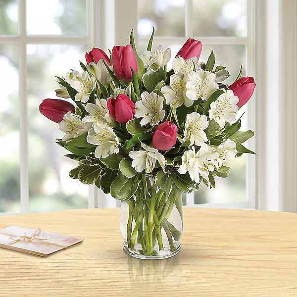 Bouquet of red and white flowers in a clear vase on a wooden table with a blurred window background.