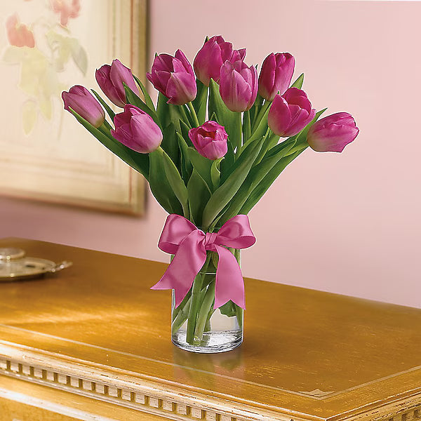Pink tulips in a glass vase with a bow on a wooden surface.