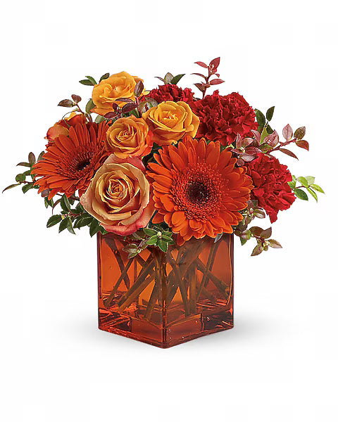 Colorful flower arrangement with orange, red, and yellow flowers in a transparent orange vase on a white background.