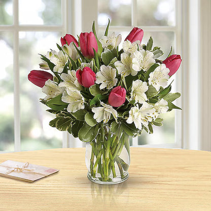 Bouquet of red and white flowers in a clear vase on a wooden table with a blurred window background.